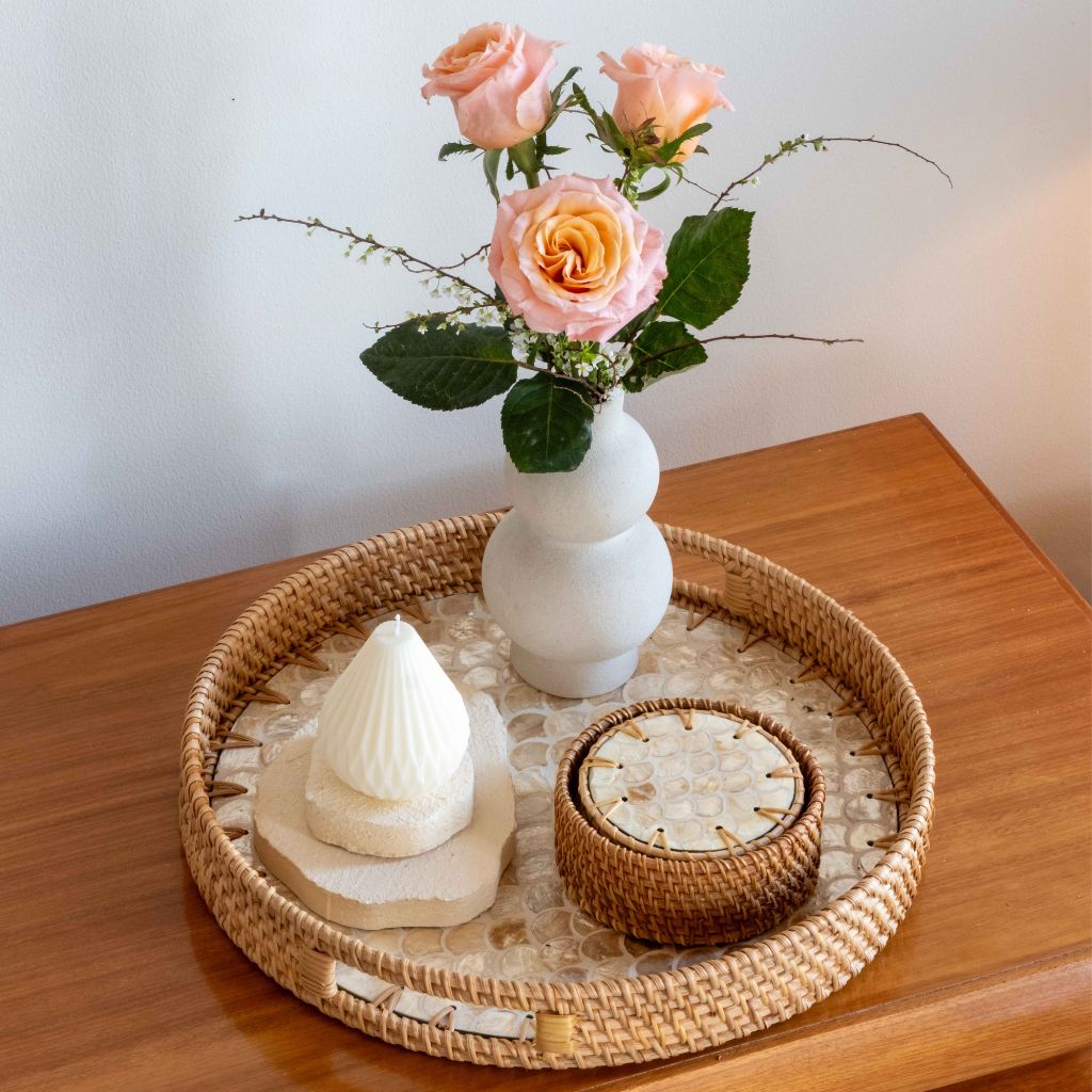 A styled home decor scene showing a round woven tray on a wooden table, holding a white ceramic vase with pink roses, a white textured candle, and a small round woven box.