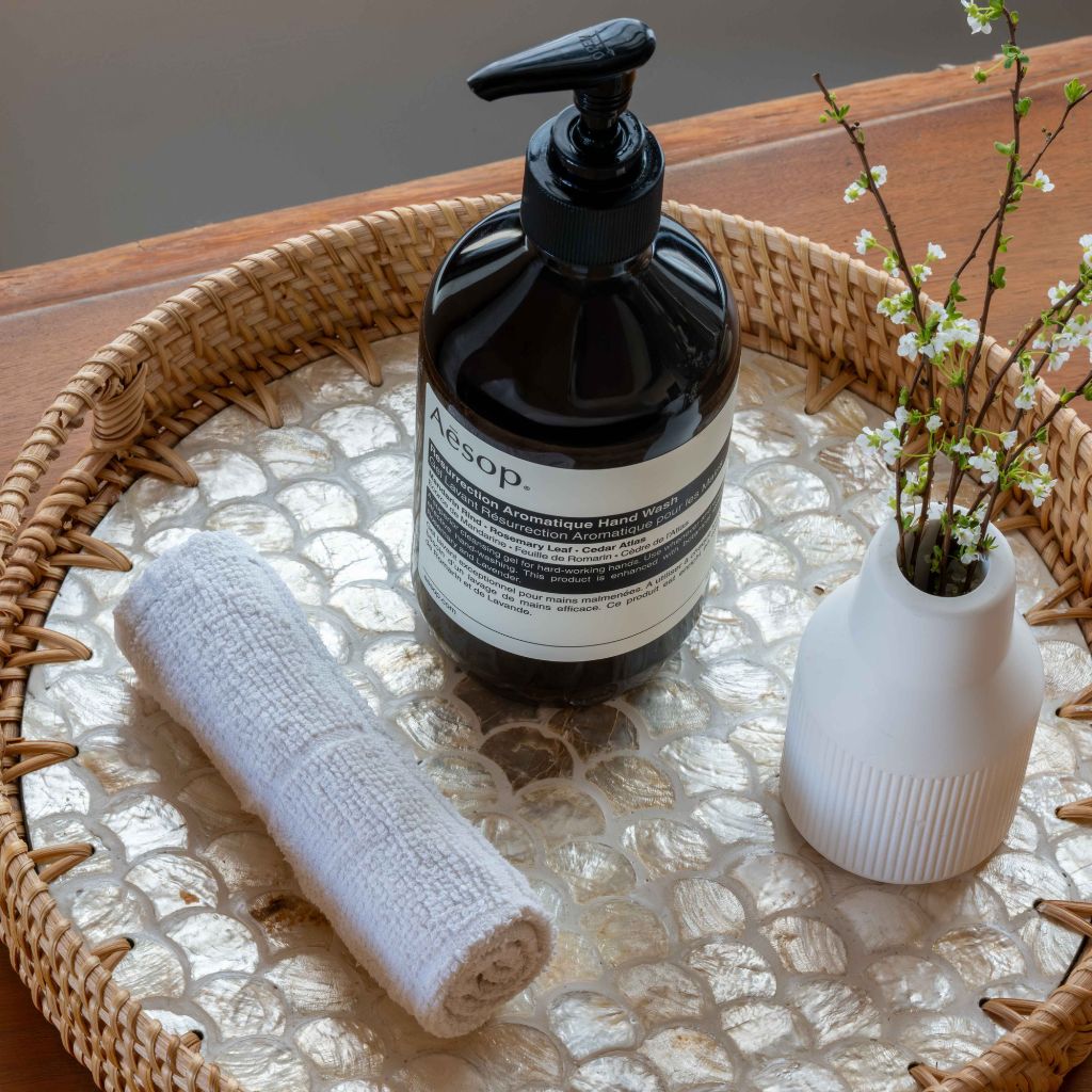 A bottle of Aesop hand soap with a black pump and white label, styled on a woven tray with a rolled white towel and a small white vase with delicate branches.